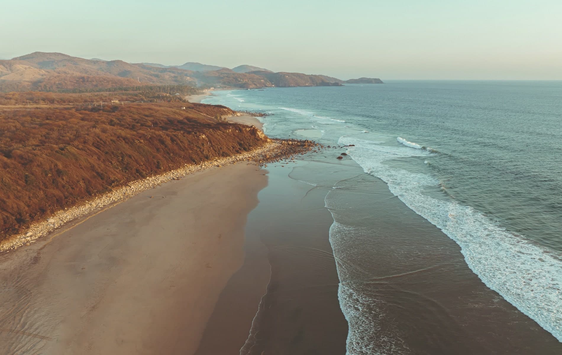 Aerial view of the Pacific coastline at MUSA — beach, ocean, and rolling mountains at golden hour